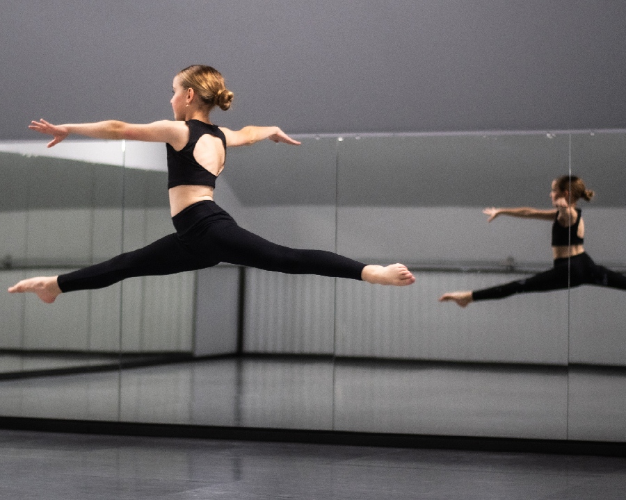 Young dancer from Tanya's Dance Company performs a high leap in a mirrored studio, showcasing technique and expression.