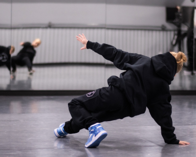 Dancer in black hoodie and sweatpants performing a breakdance move in a mirrored studio.